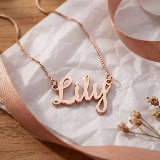 A rose gold plated name necklace reading Lily on crumpled tissue beside a satin ribbon