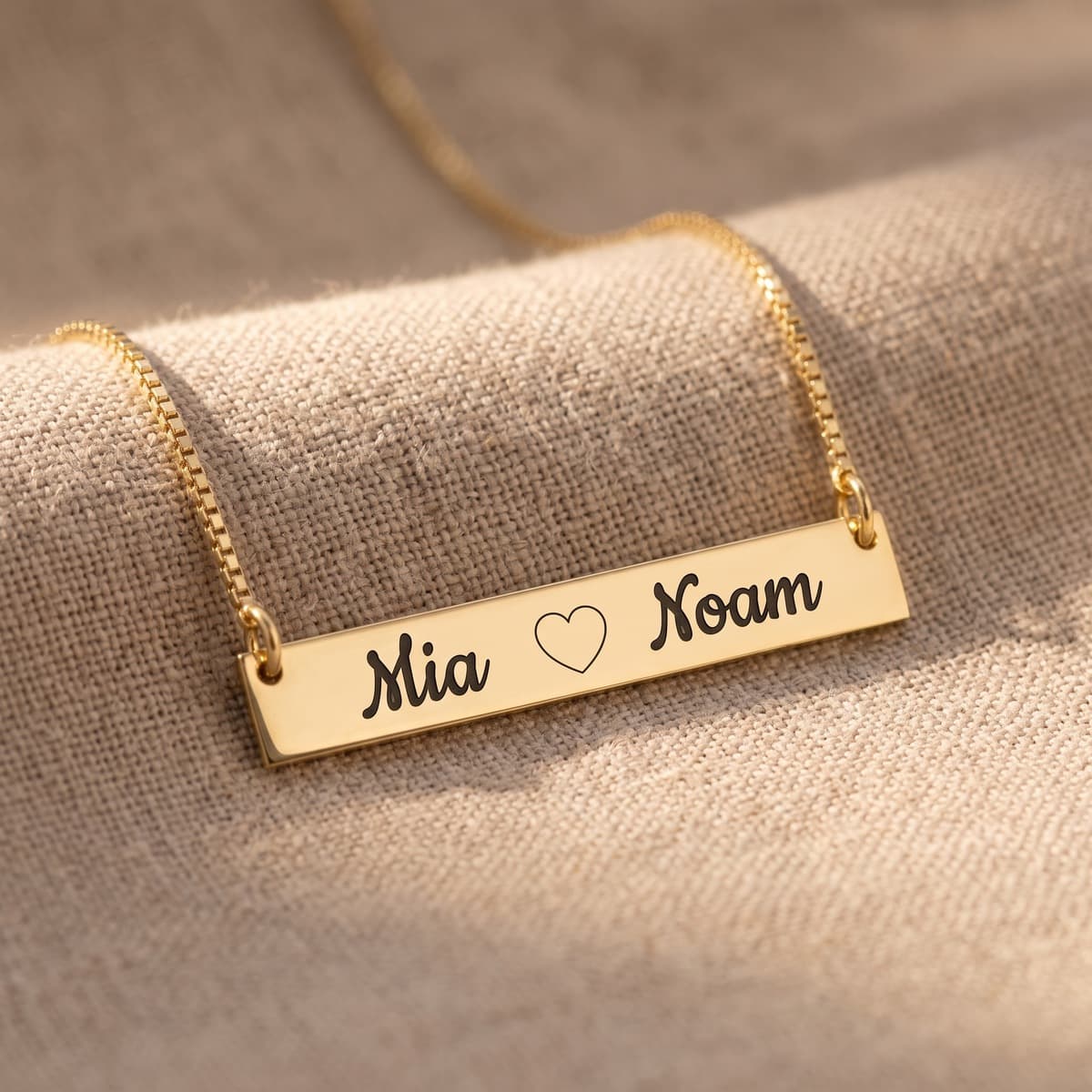 A gold-plated bar necklace engraved “Mia ♡ Noam” on linen