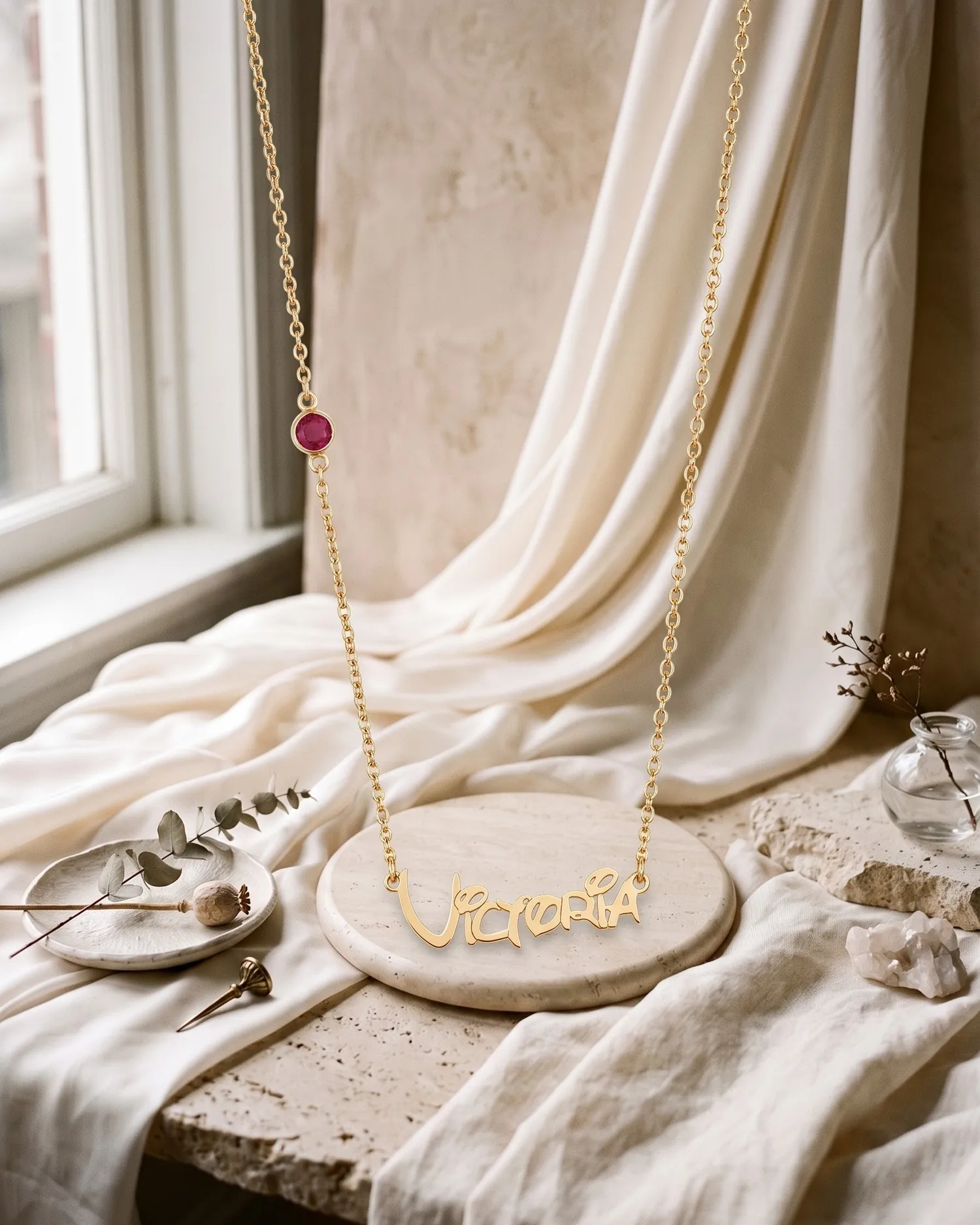 A gold plated name necklace reading “Victoria” with a birthstone, on a stone plate with ivory silk
