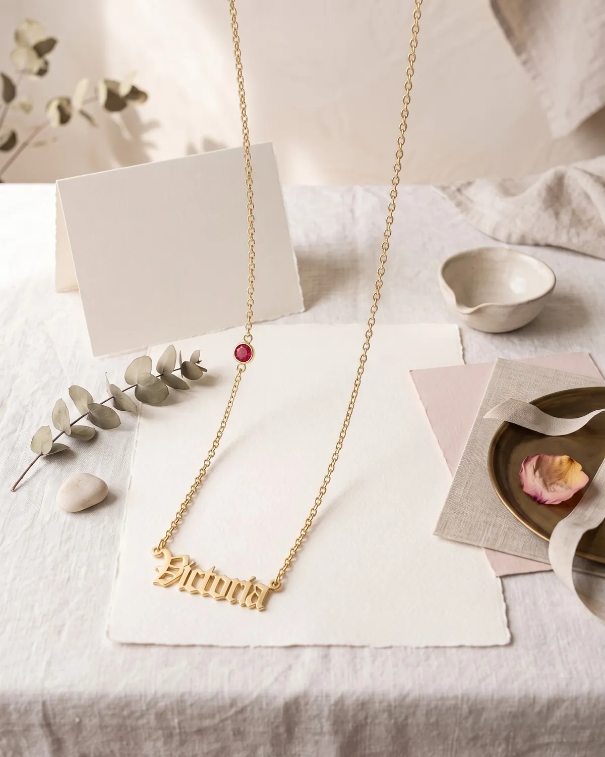 A gold plated Old English name necklace reading “Victoria” with a birthstone, on cream paper