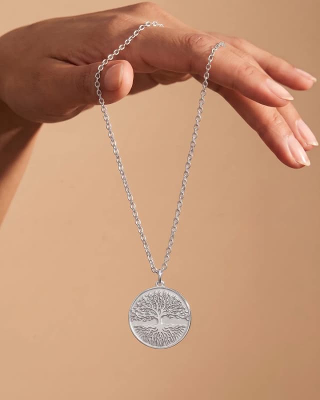 A 925 sterling silver tree of life disc necklace hanging from a hand on a warm sand ground, a photograph made in the AI Studio