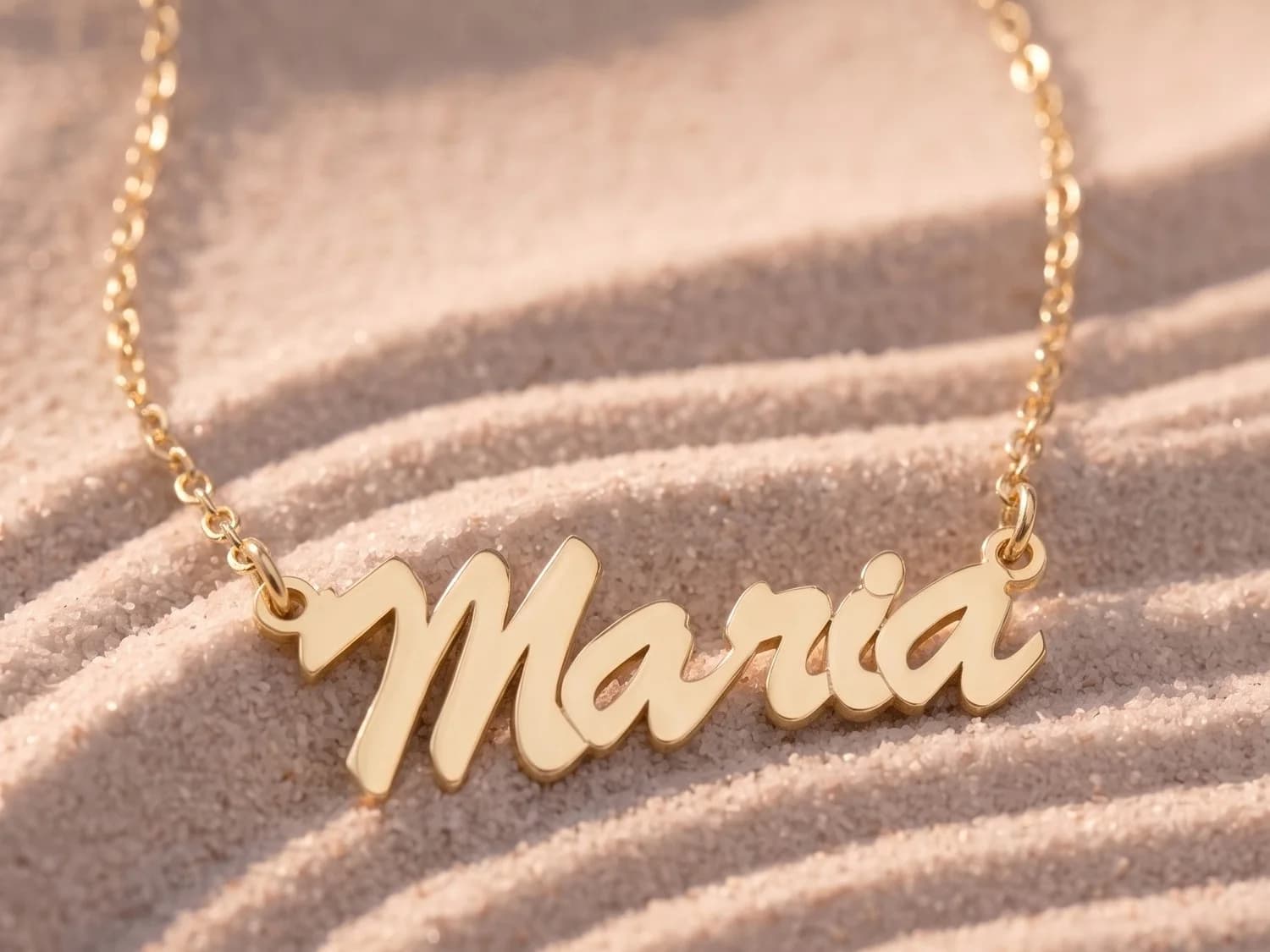 A gold plated name necklace reading “Maria” on a cable chain, laid on rippled sand