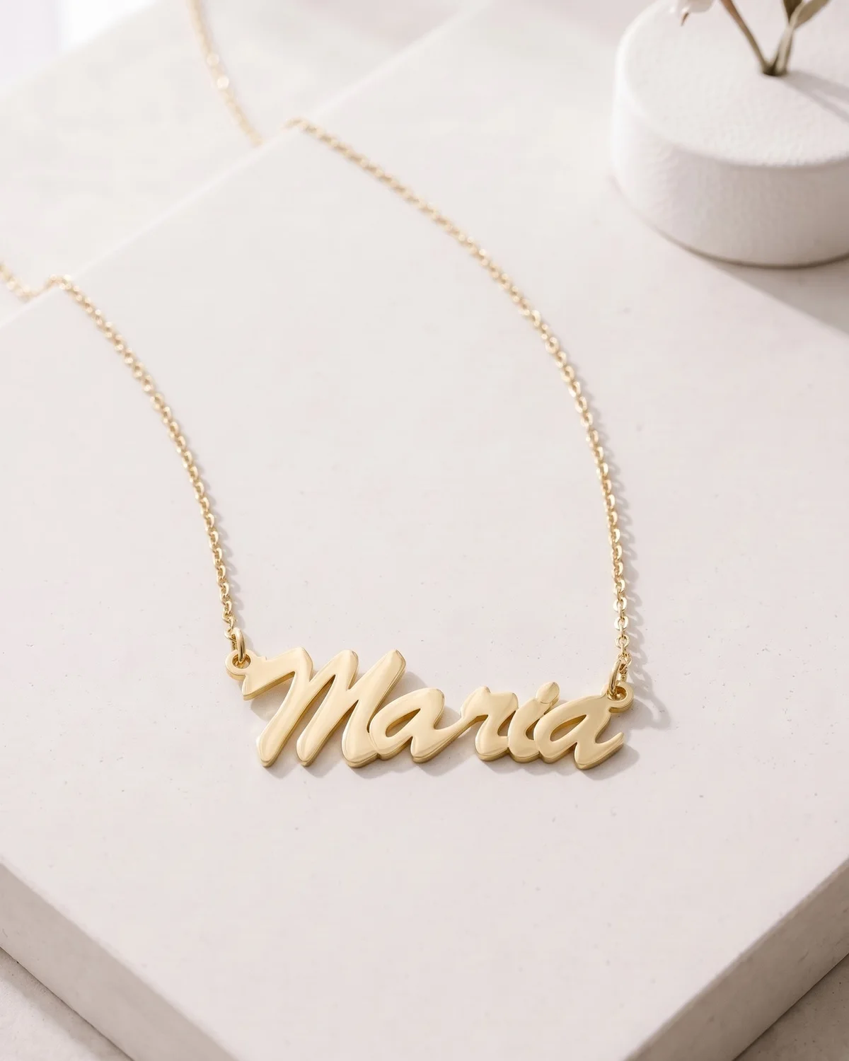 A gold plated name necklace reading “Maria” on a cable chain, on white paper by a small vase