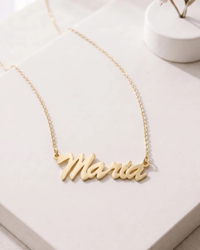 A gold plated name necklace reading “Maria” on a cable chain, on white paper by a small vase